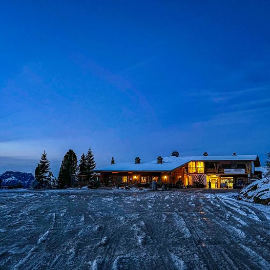 Ein gemütliches Chalet, das bei Nacht beleuchtet ist, umgeben von Schnee und Kiefern, mit einer Person, die das Gebäude betritt.