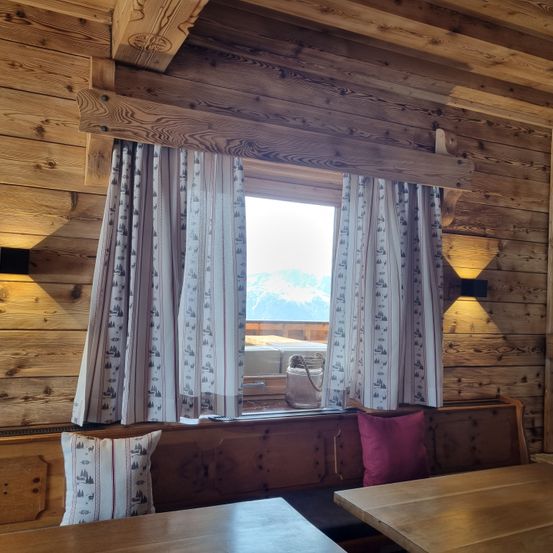 Ein gemütliches Blockhütten-Interieur mit Holzwänden, einem Fenster mit Vorhängen und einer Bank mit Kissen. Der Raum bietet einen Blick auf die Berge.