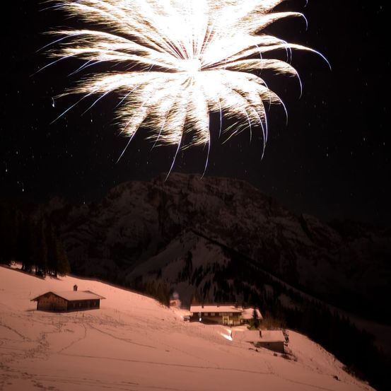 Ein Feuerwerk explodiert in der Nacht über einer verschneiten Landschaft mit einem Haus und einem Berg im Hintergrund.