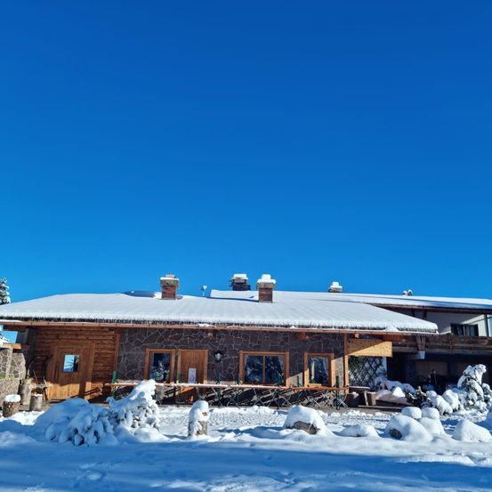 Ein Chalet mit Holzwänden und schneebedecktem Dach steht unter einem klaren blauen Himmel. Es gibt schneebedeckte Außentische und Stühle sowie Schneehaufen darum herum.