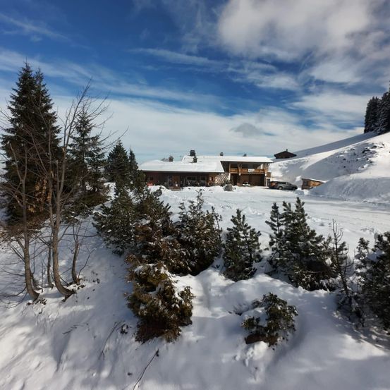 Ein großes Gebäude mit weißem Dach ist von schneebedeckten Bäumen und Büschen umgeben. Das Gebäude befindet sich auf einem verschneiten Berg mit einem klaren blauen Himmel.