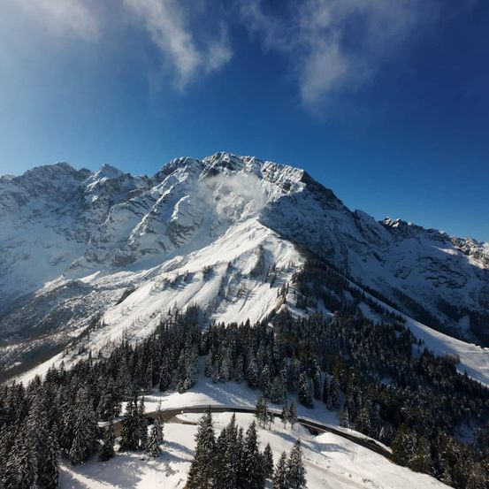 Ein verschneiter Berggipfel mit Kiefern und einer kurvenreichen Straße unter einem blauen Himmel mit Wolken.