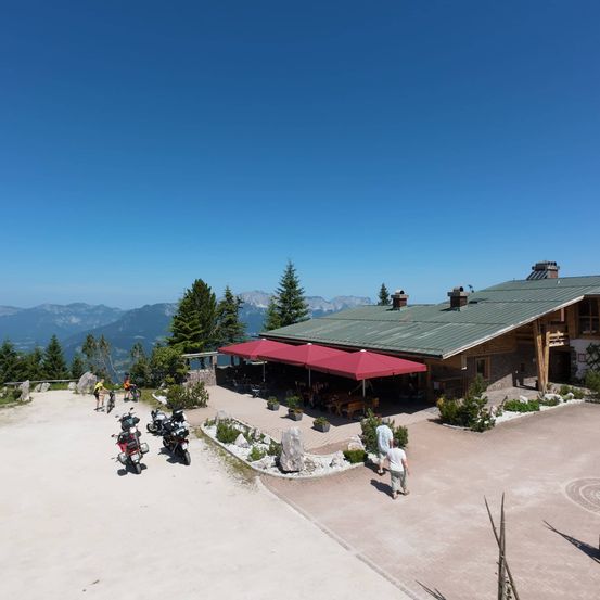 Eine Luftaufnahme eines Berggasthauses mit roten Sonnenschirmen, mehreren geparkten Motorrädern und spazieren gehenden Menschen vor dem Hintergrund von Bäumen und Bergen.