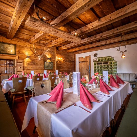 Ein Essbereich mit Holzbalken und rustikalem Dekor, mit langen Tischen, die mit weißen Tischdecken, rosa Servietten und Besteck gedeckt sind. Der Raum hat Holzstühle, Geweihe an den Wänden und eine Holztür.