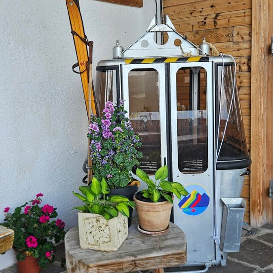 Eine Skiliftkabine mit Glasfenstern und einer Glastür steht draußen. Sie befindet sich in der Nähe einer weißen Wand. Davor stehen Topfpflanzen und Blumen.