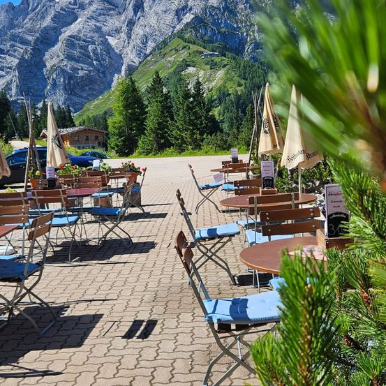 Eine Terrasse eines Cafés in den Bergen mit Tischen und Stühlen, umgeben von Bäumen und Bergen im Hintergrund.