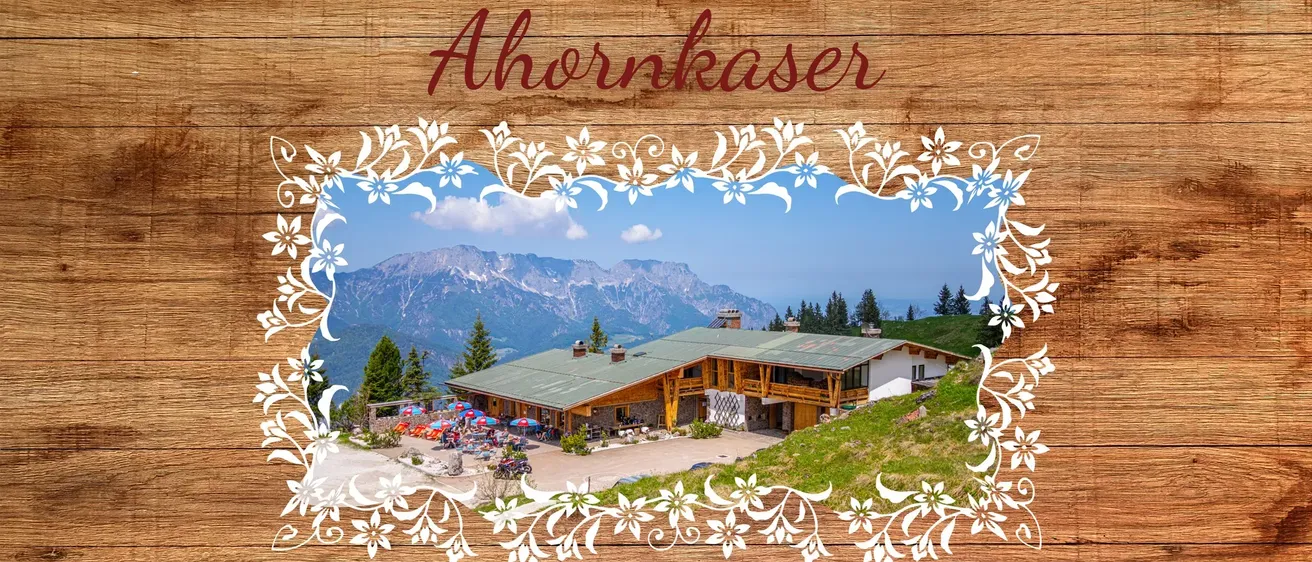 Ein Poster mit einem hölzernen Hintergrund zeigt ein Haus in den Bergen mit dem Text 'Servus und Herzlich Willkommen bei uns am Ahornkaser'. Im Vordergrund ist ein großes Gebäude mit einem grünen Dach und einer weißen Außenfassade zu sehen. Mehrere Personen stehen davor, einige unter Regenschirmen. Im Hintergrund befinden sich Berge und Bäume.