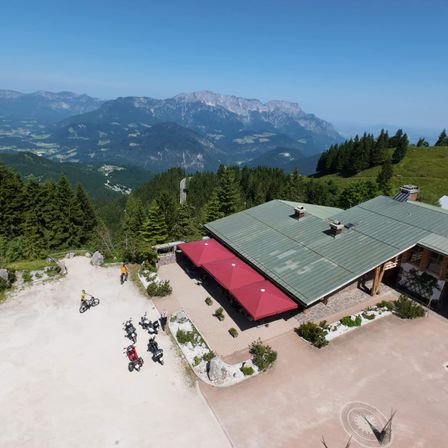 Eine Luftaufnahme eines Gebäudes mit grünem Dach, roten Markisen und mehreren geparkten Motorrädern davor. Die Umgebung ist von üppigen Bäumen umgeben und im Hintergrund befinden sich Berge.