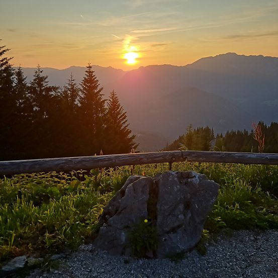 Sonnenuntergang vom Gipfel eines Berges aus gesehen, mit einem großen Felsbrocken im Vordergrund und einem Holzzaun. Der Himmel ist mit warmen Orange- und Rosatönen erfüllt.