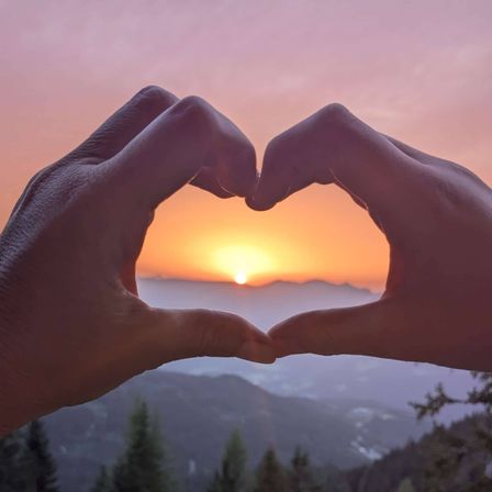 Zwei Hände bilden ein Herz in der Dämmerung mit Bergen in der Ferne.