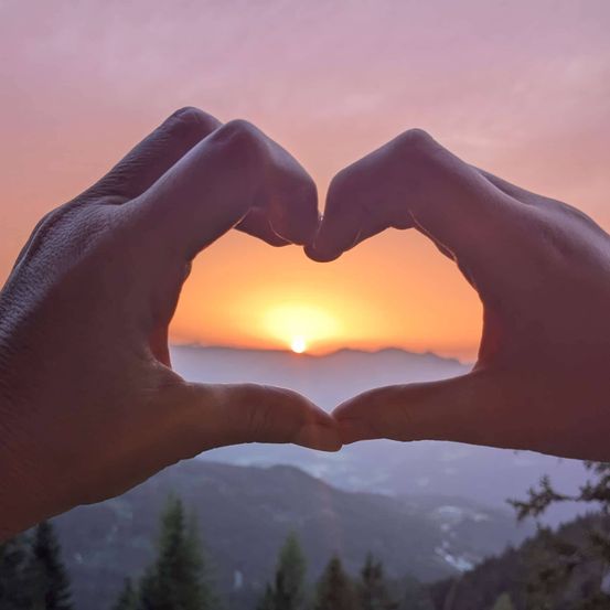 Zwei Hände bilden ein Herz in der Dämmerung mit Bergen in der Ferne.