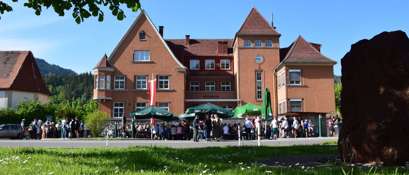 Ein großes braunes Gebäude mit vielen Fenstern und einer Uhr an der Seite. Es gibt Menschen, die draußen sitzen und stehen, unter grünen Sonnenschirmen. Davor befindet sich eine Rasenfläche.