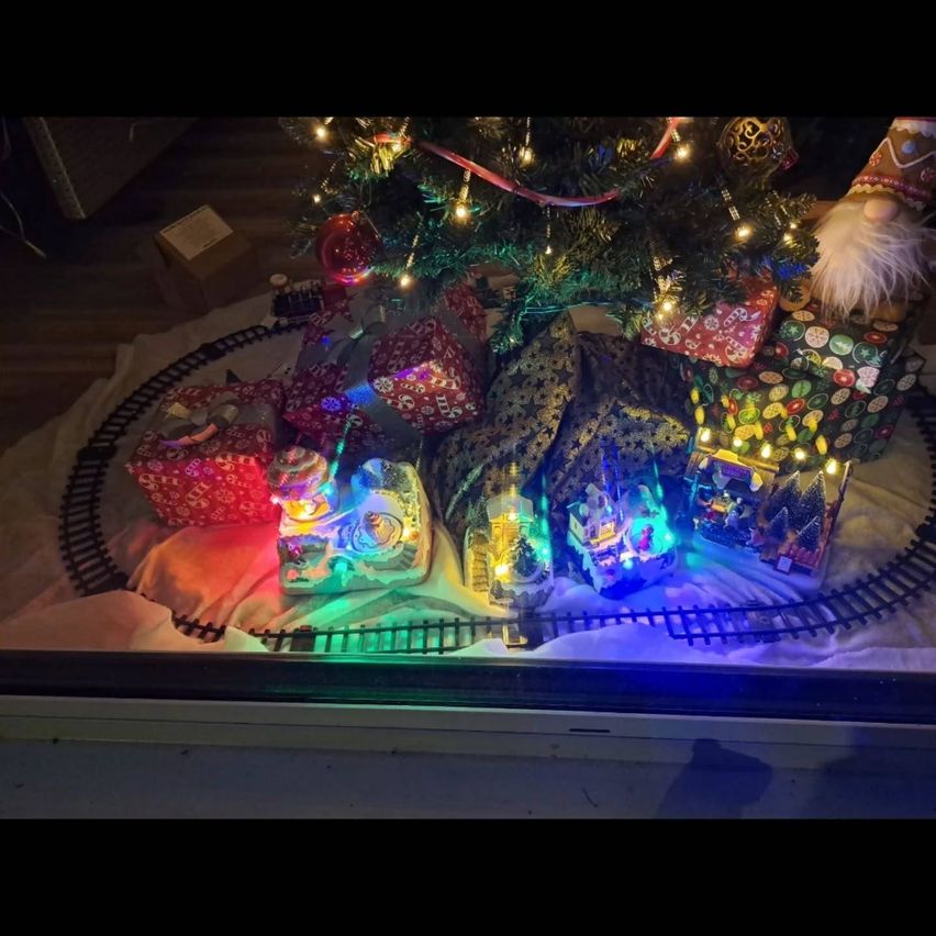 Ein Weihnachtsbaum mit bunten Lichtern und verpackten Geschenken steht auf einer Miniatur-Eisenbahnstrecke, umgeben von einer festlichen Umgebung.