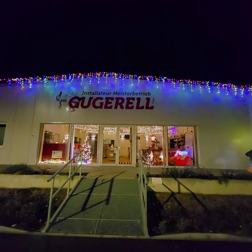 Ein Ladengeschäft bei Nacht mit festlichen Lichtern, einem Weihnachtsbaum und Dekorationen. Das Schild zeigt 'Gugerell' mit einem Logo.