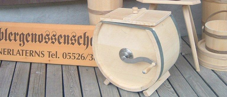 Eine hölzerne Presse steht auf einer Holzterrasse mit Holzeimern und einem kleinen Stuhl mit Blumenvase. Dahinter befindet sich eine Holzwand mit einem Schild.