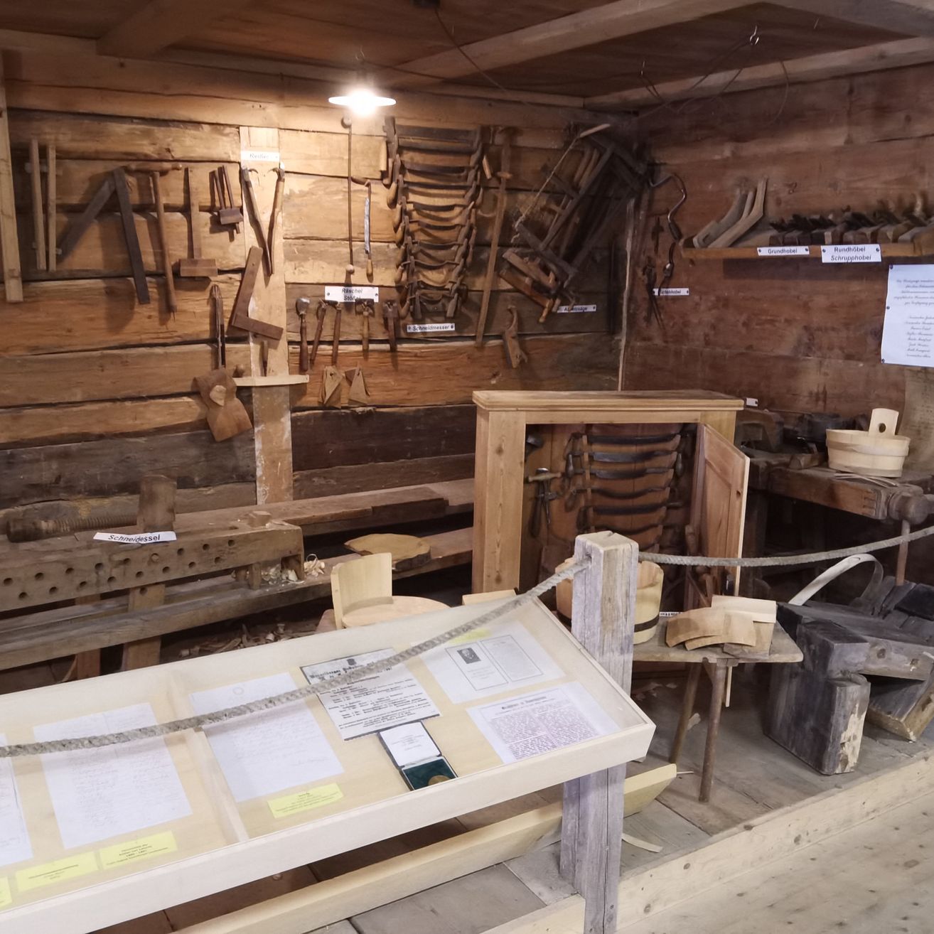 Ein rustikaler Werkstattraum mit Holzwänden, hängenden Werkzeugen und einem Schreibtisch. Papiere auf einem Ausstellungsstand. Eine Leiter in der Nähe.