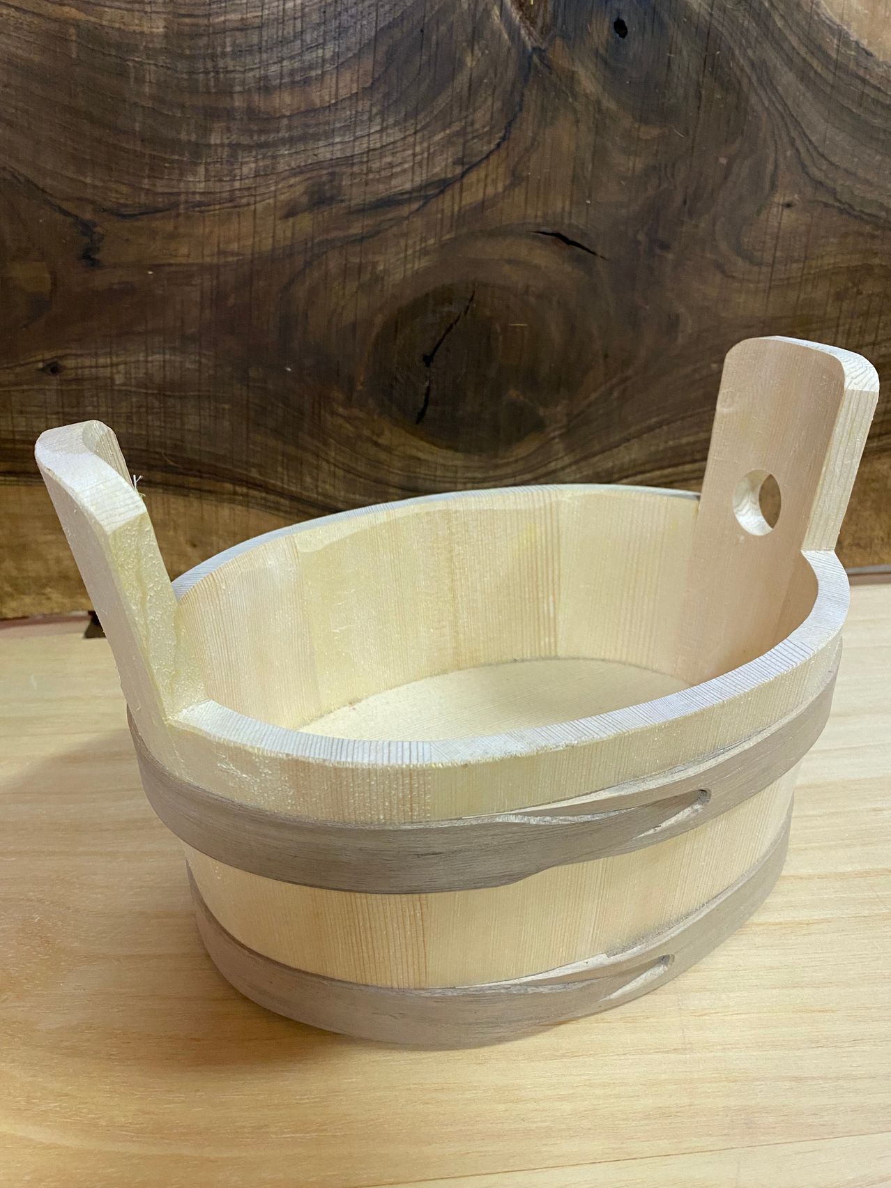 Ein kleiner Holzeimer mit zwei Griffen und einem Loch an der Seite, auf einer Holzoberfläche platziert.