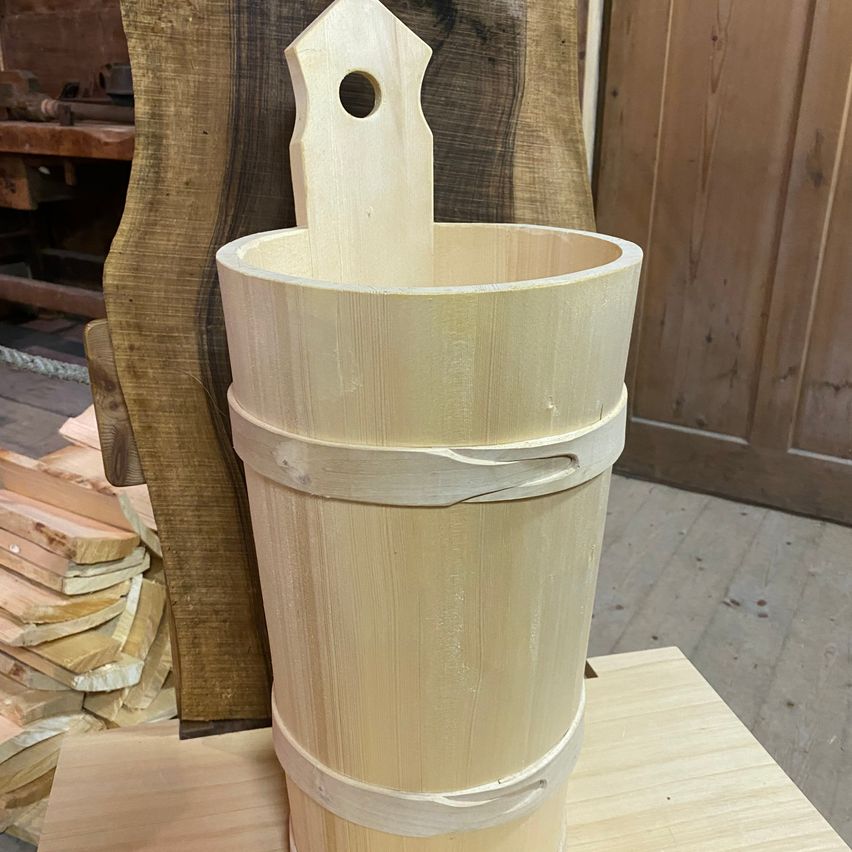 Ein Holzeimer mit Griff steht auf einer Holzoberfläche, mit einer hölzernen Struktur und Holzbrettern im Hintergrund.