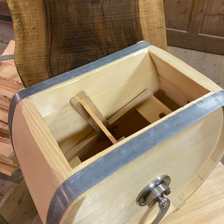 Ein hölzerner Fass mit Metallrand, geöffnet, zeigt einen internen Mechanismus und einen silbernen Griff. Dahinter befinden sich Holzbretter und eine Holztür.
