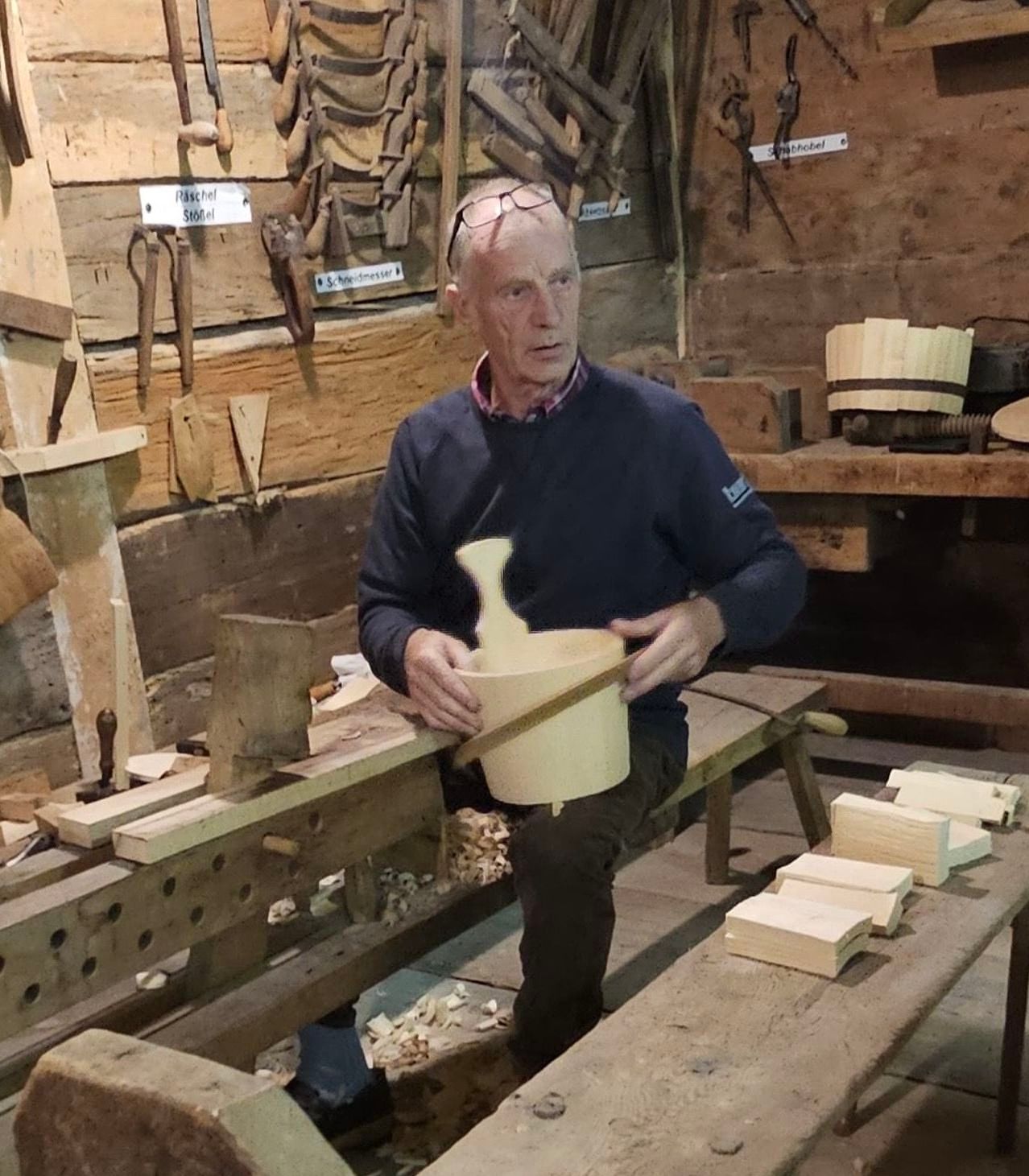 Ein älterer Mann mit Brille und blauem Pullover sitzt auf einer Bank in einer Werkstatt und hält ein Holzobjekt. Hinter ihm befinden sich Holzwerkzeuge und eine Holzwand mit einem Schild.