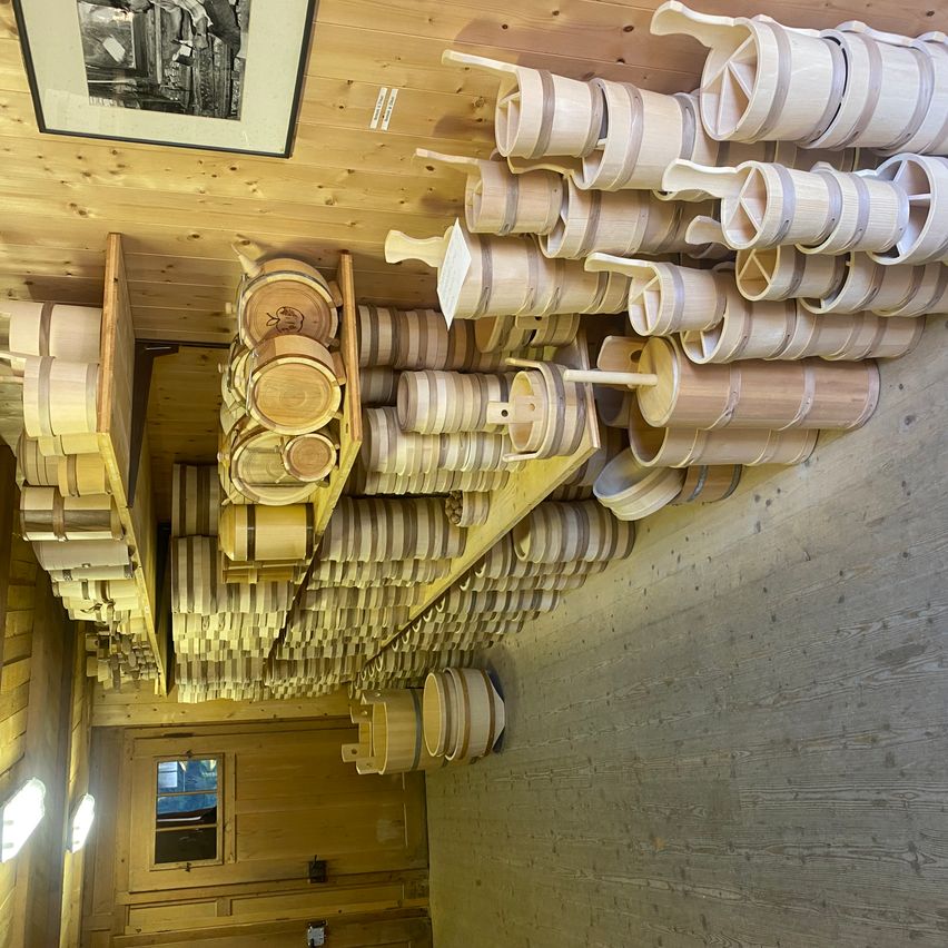 A wooden room with shelves of wooden buckets and barrels stacked in different sizes and shapes. The room has wooden walls and floor, a window, and a picture frame.