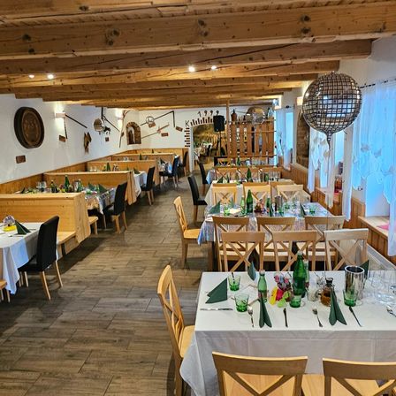 Ein Restaurant mit Holztischen, Stühlen und einer Holzdecke. Die Tische sind mit Flaschen, Gläsern und Besteck gedeckt. Die Wände sind mit einer Uhr, einem Holzregal und anderen Gegenständen dekoriert. Der Boden ist aus Holz.