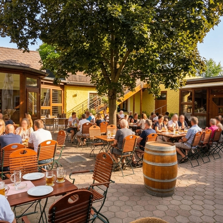 Ein sonniger Tag in einem geschäftigen Außenrestaurant. Die Leute sitzen an Holztischen unter einem großen Baum und genießen Getränke und Mahlzeiten. Das Lokal hat gelbe Wände und Glasfenster.