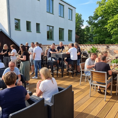 Eine Gruppe von Menschen sitzt an Tischen auf einer Terrasse im Freien, einige stehen und halten Getränke. Im Hintergrund sind Bäume und ein Gebäude mit Glasfenstern zu sehen.
