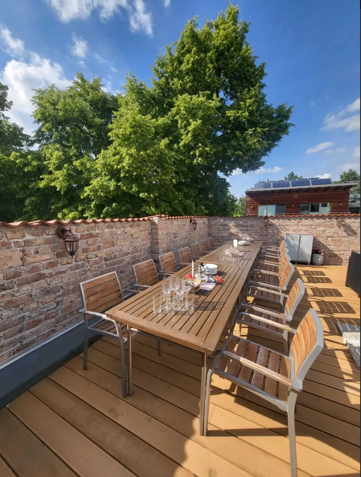 Ein langer Holztisch auf einer Holzterrasse mit mehreren Stühlen. Eine Ziegelmauer und ein Gebäude mit Solarmodulen im Hintergrund.