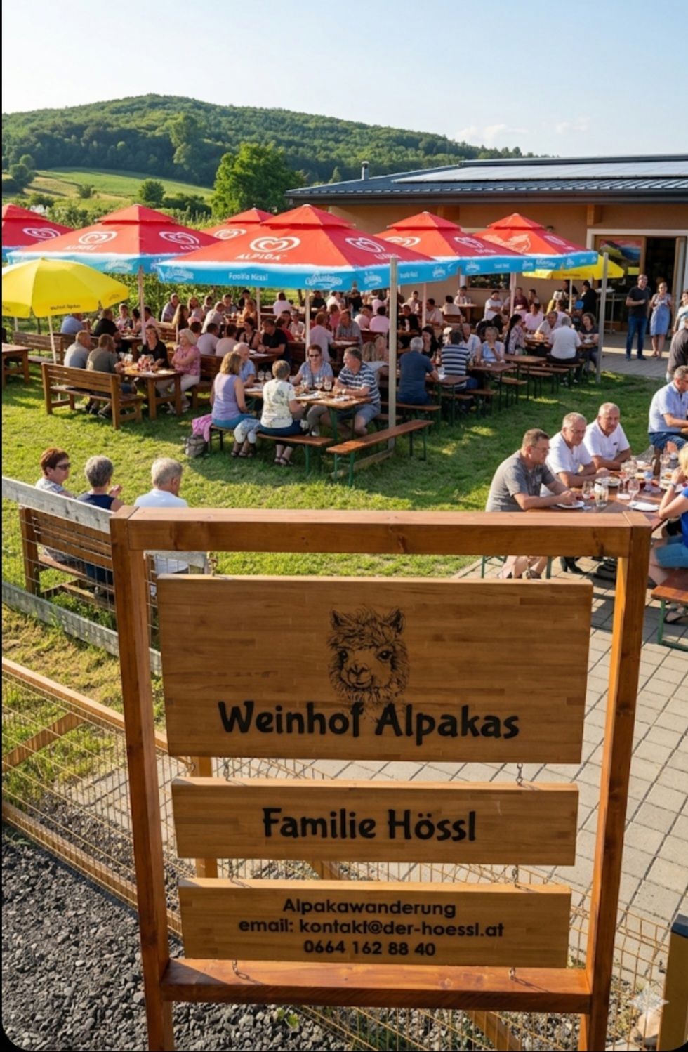 Ein Outdoor-Restaurant mit Tischen und Bänken unter Schirmen, mit sitzenden und stehenden Personen. Ein Holzschild mit einem Alpaka und dem Text 'Weinhof Alpakas Familie Hossl' ist im Vordergrund.