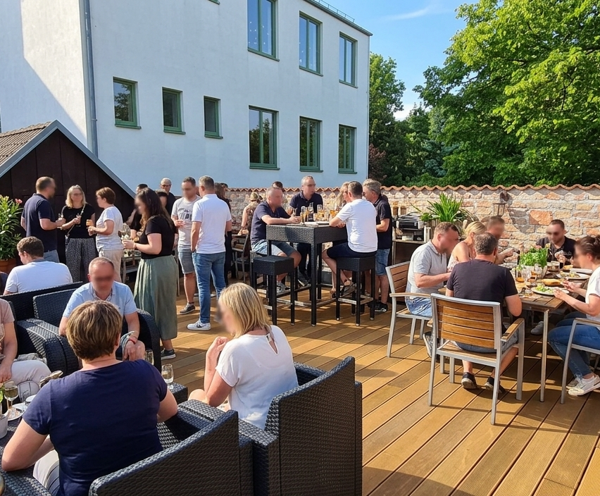 Mehrere Menschen sitzen auf Stühlen und Tischen in einem Restaurant im Freien. Einige stehen und halten Gläser. Das Restaurant hat einen Holzboden und eine Wand mit Glasfenstern.