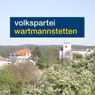 Ein landschaftlicher Blick auf ein Dorf mit blühenden Bäumen und Häusern, mit einem blauen Schild im Vordergrund, das die Worte 'Volkspartei Wartmannstetten' trägt.