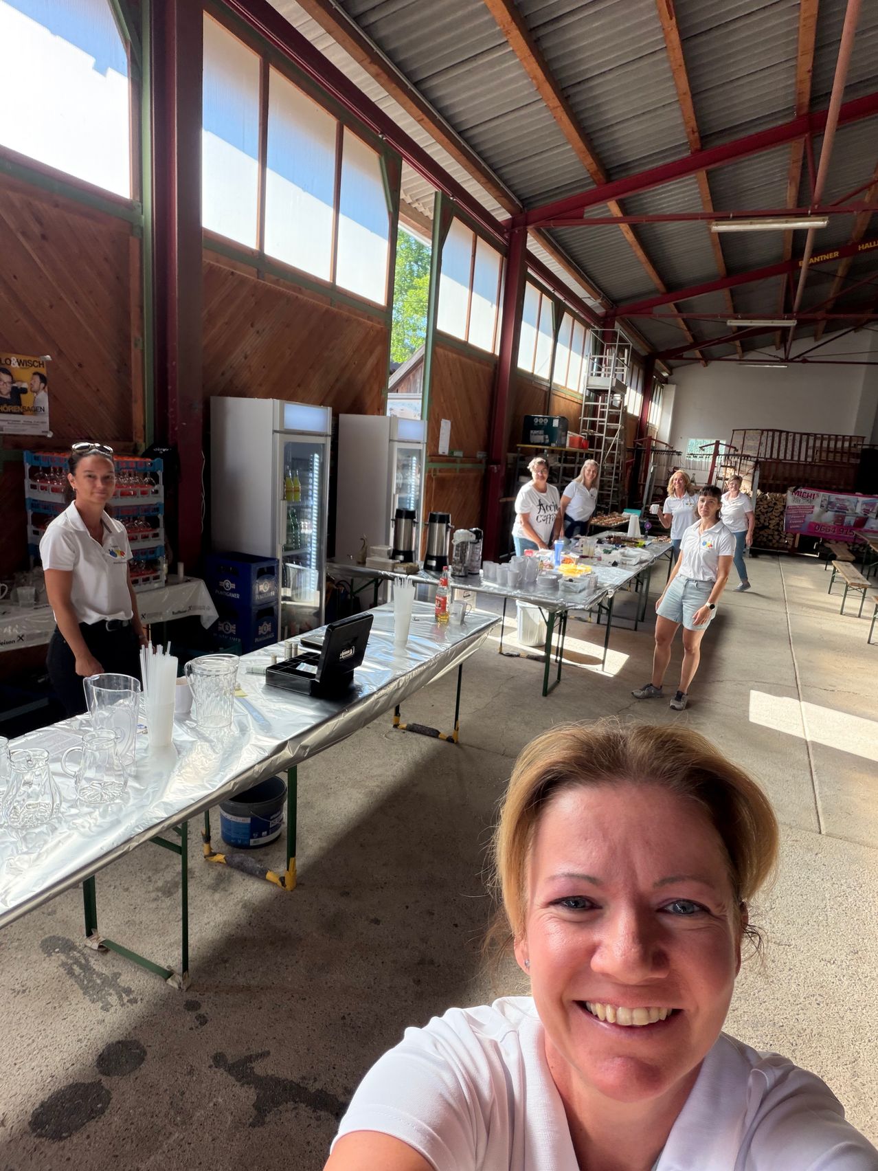A group of people are setting up a table in a large room with wooden walls and large windows. They are wearing white shirts and shorts. There are several items on the table, including cups, a coffee machine, and a refrigerator. A woman is standing in front of the table, smiling.