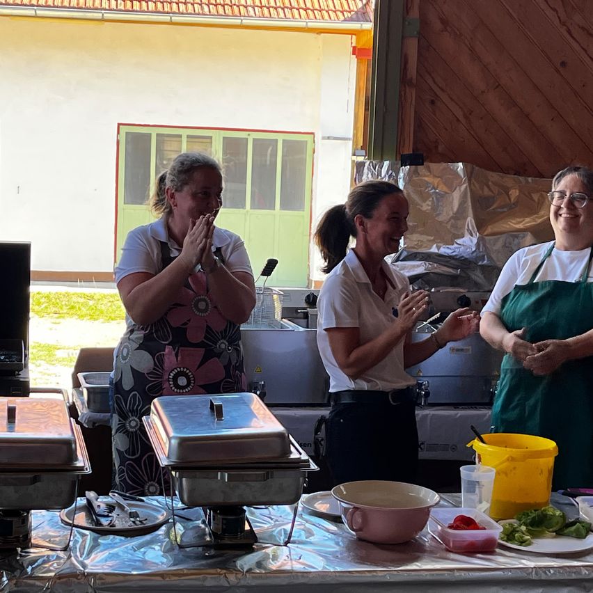 Three women in aprons stand behind a table with food prep items, smiling and clapping. The background shows a building with green doors.