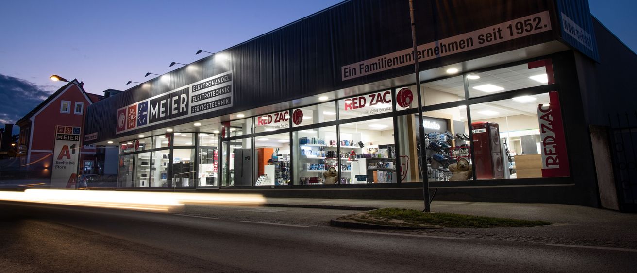 Ein Geschäft mit einem RED ZAC-Schild und einer Straßenlampe davor, beleuchtet in der Dämmerung, zeigt eine Vielzahl von Artikeln im Inneren.