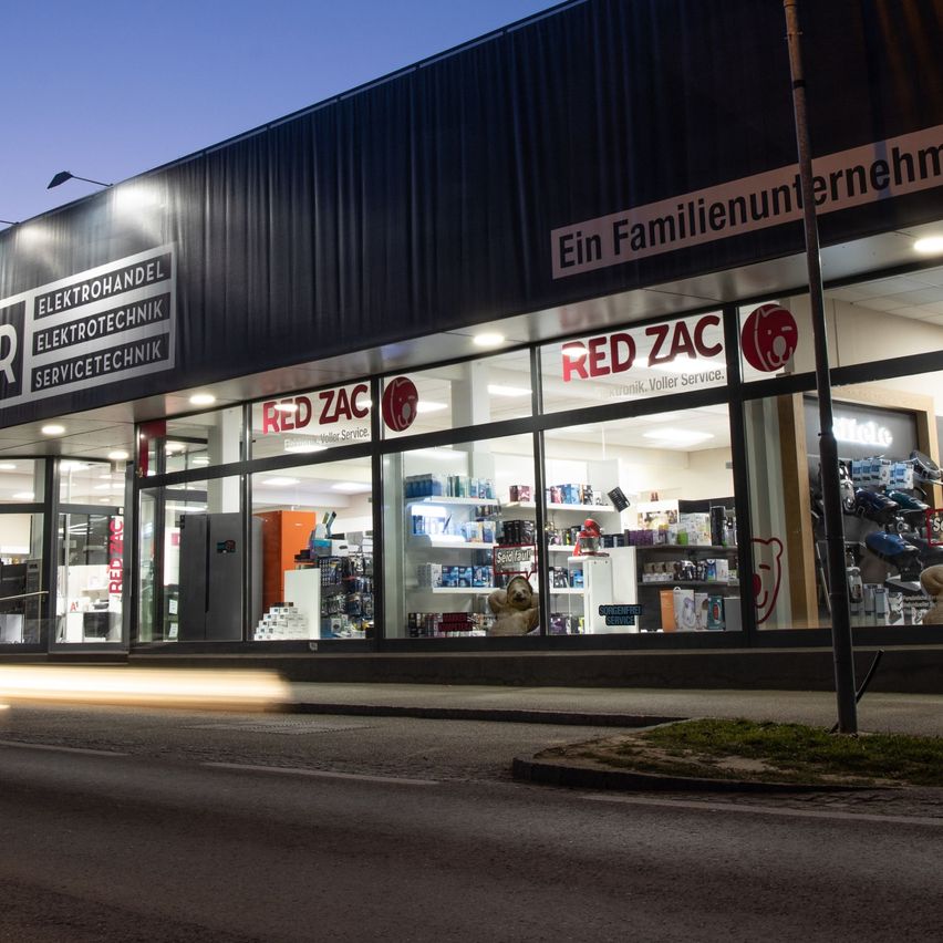 Ein Geschäft mit einem RED ZAC-Schild und einer Straßenlampe davor, beleuchtet in der Dämmerung, zeigt eine Vielzahl von Artikeln im Inneren.