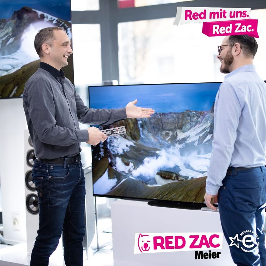 Zwei Männer stehen vor einem großen Fernsehbildschirm. Ein Mann hält eine Fernbedienung in der Hand und zeigt auf den Bildschirm, während der andere Mann zusieht. Dahinter befindet sich eine Glaswand mit einem Bild einer Berglandschaft.