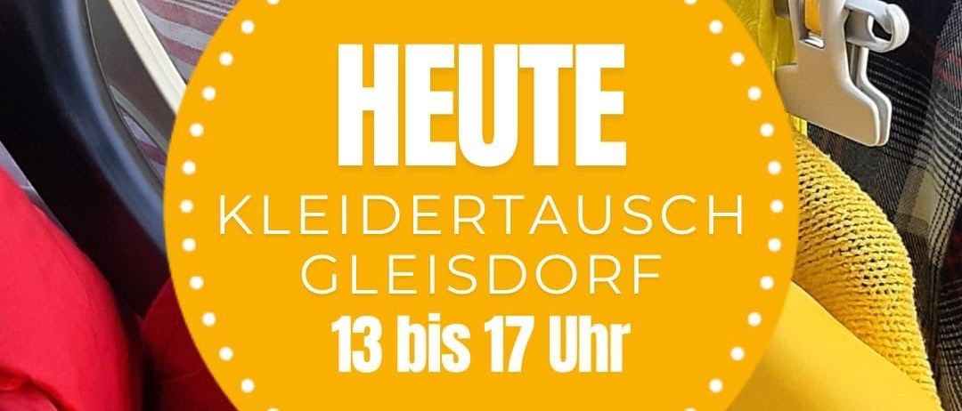 Ein gelber Kreisschild mit weißem Text sagt Heute Kleidertausch Gleisdorf 13 bis 17 Uhr über einem Kleiderständer mit farbenfrohen Klamotten.