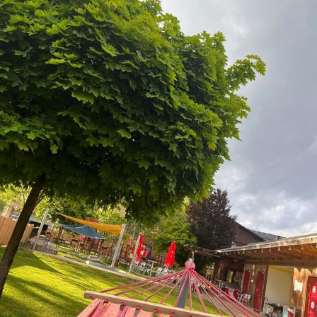 Ein sonniger Tag in einem Park mit einem großen Baum, einer Hängematte und einem Gebäude mit roten Türen und Sonnenschirmen.