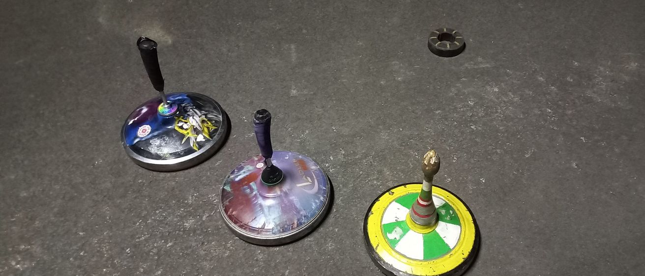 Mehrere Objekte liegen auf einem Betonboden. Es gibt einen lilafarbenen Top, ein kreisförmiges gelb-grünes Objekt und einen kleinen schwarzen Ring.