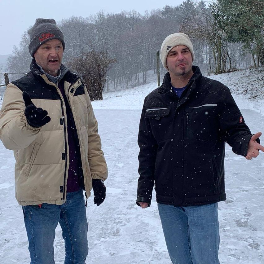 Zwei Männer stehen draußen im Schnee und tragen Winterjacken und Mützen. Einer gestikuliert, während der andere ihn ansieht. Im Hintergrund sind Bäume und ein Zaun zu sehen.