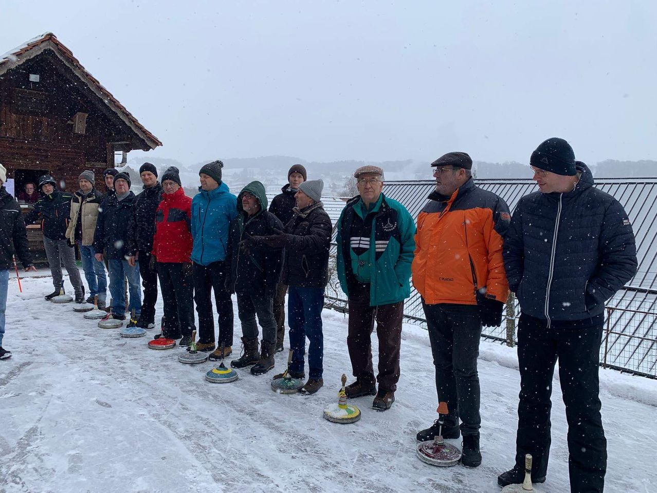 Eine Gruppe von Männern steht in einer Reihe auf einem verschneiten Feld und trägt Winterkleidung. Sie bereiten sich auf eine Partie Curling vor. Der Boden ist mit Schnee bedeckt.