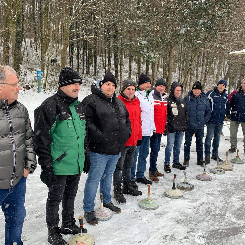 Eine Gruppe von Männern in Winterjacken und Mützen steht im Schnee und bereitet sich auf ein Curling-Match vor. Sie stehen vor hölzernen Curling-Zielen auf einem verschneiten Feld.