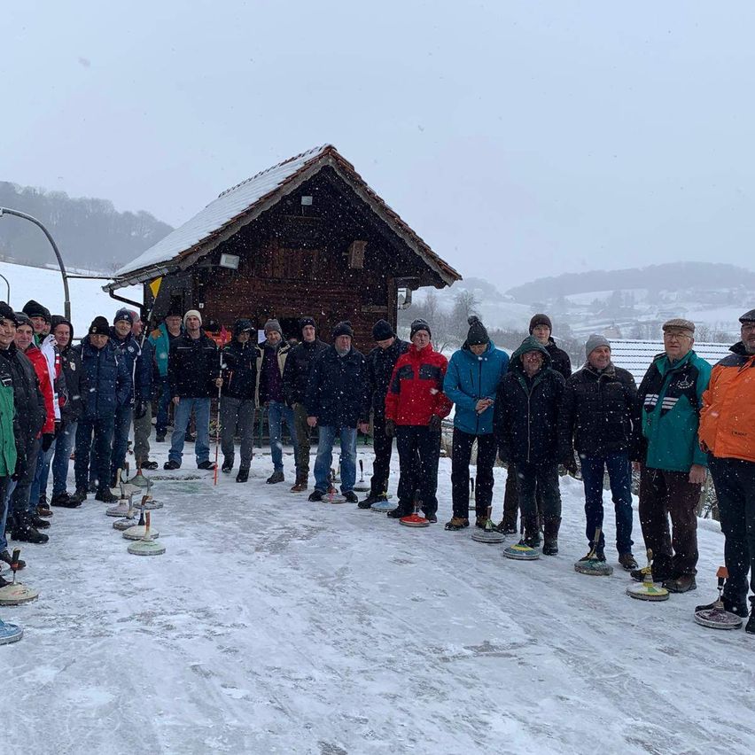 Eine Gruppe von Menschen, die warm gekleidet sind, steht in einem verschneiten Feld und hält Curling-Broomsticks. Hinter ihnen steht ein hölzernes Chalet mit Schnee auf dem Dach in der Nähe eines Straßenlaternenmastes. In der Ferne sind Berge unter einem bewölkten Himmel sichtbar.