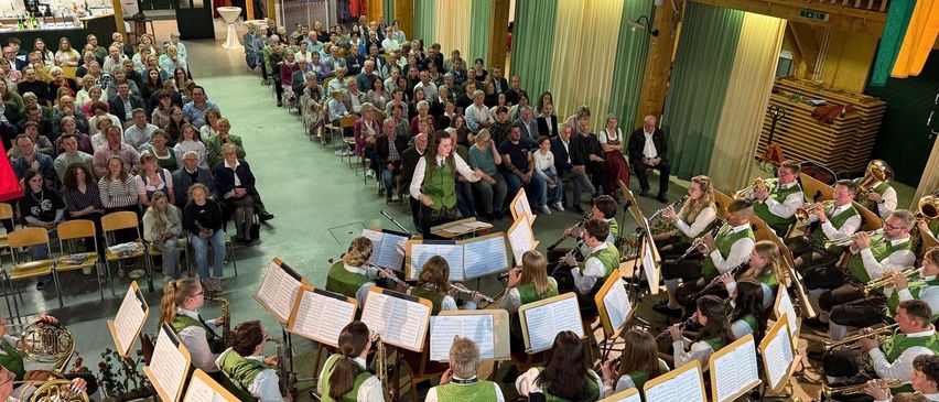Ein großer Innensaal mit Holzbalken und grünen Vorhängen. Ein Blasorchester spielt, wobei die Musiker verschiedene Instrumente spielen. Das Publikum sitzt in Reihen und beobachtet die Aufführung. Der Saal hat auch eine Galerie mit Zuschauern.