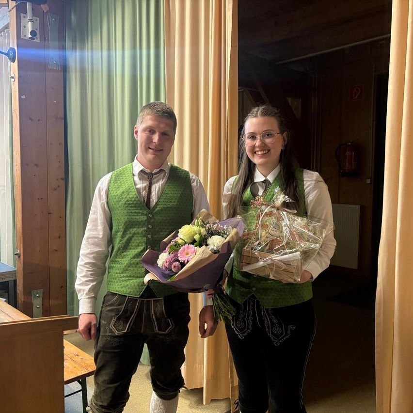 Ein Mann und eine Frau stehen zusammen und halten Blumen und Geschenke in den Händen. Der Mann trägt eine grüne Weste und die Frau trägt eine Brille.