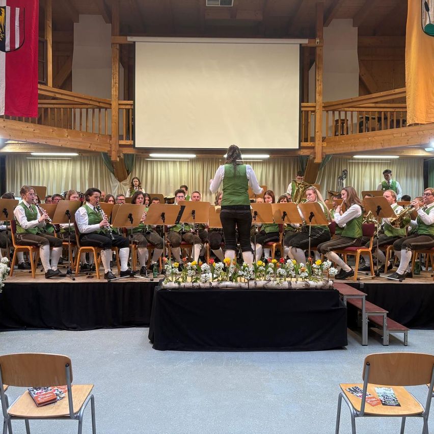 Eine Band spielt auf der Bühne mit verschiedenen Instrumenten. Eine Person steht vorne, umgeben von Blumen. Im Hintergrund hängen zwei Fahnen, und das Publikum sitzt in der vorderen Reihe.