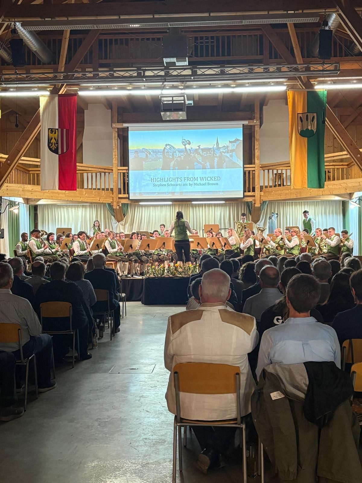 Eine Gruppe von Menschen sitzt in Stühlen und beobachtet eine Band auf der Bühne mit einer Projektionsleinwand dahinter.