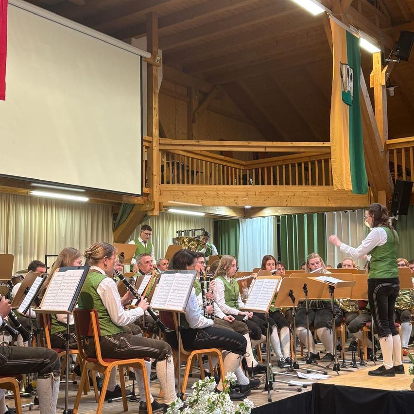 Eine Gruppe von Musikern in grünen Westen und schwarzen Hosen spielt verschiedene Instrumente auf der Bühne, angeführt von einer Frau in einem grünen West. Sie stehen vor einem weißen Vorhang, mit einer Projektionsleinwand an der Wand.