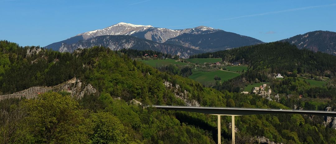 Ein Gebirgszug mit schneebedeckten Gipfeln steht unter einem klaren blauen Himmel. Darunter ist ein mit Grün bewachsenes Tal mit Häusern zu sehen, mit einer Brücke im Vordergrund.
